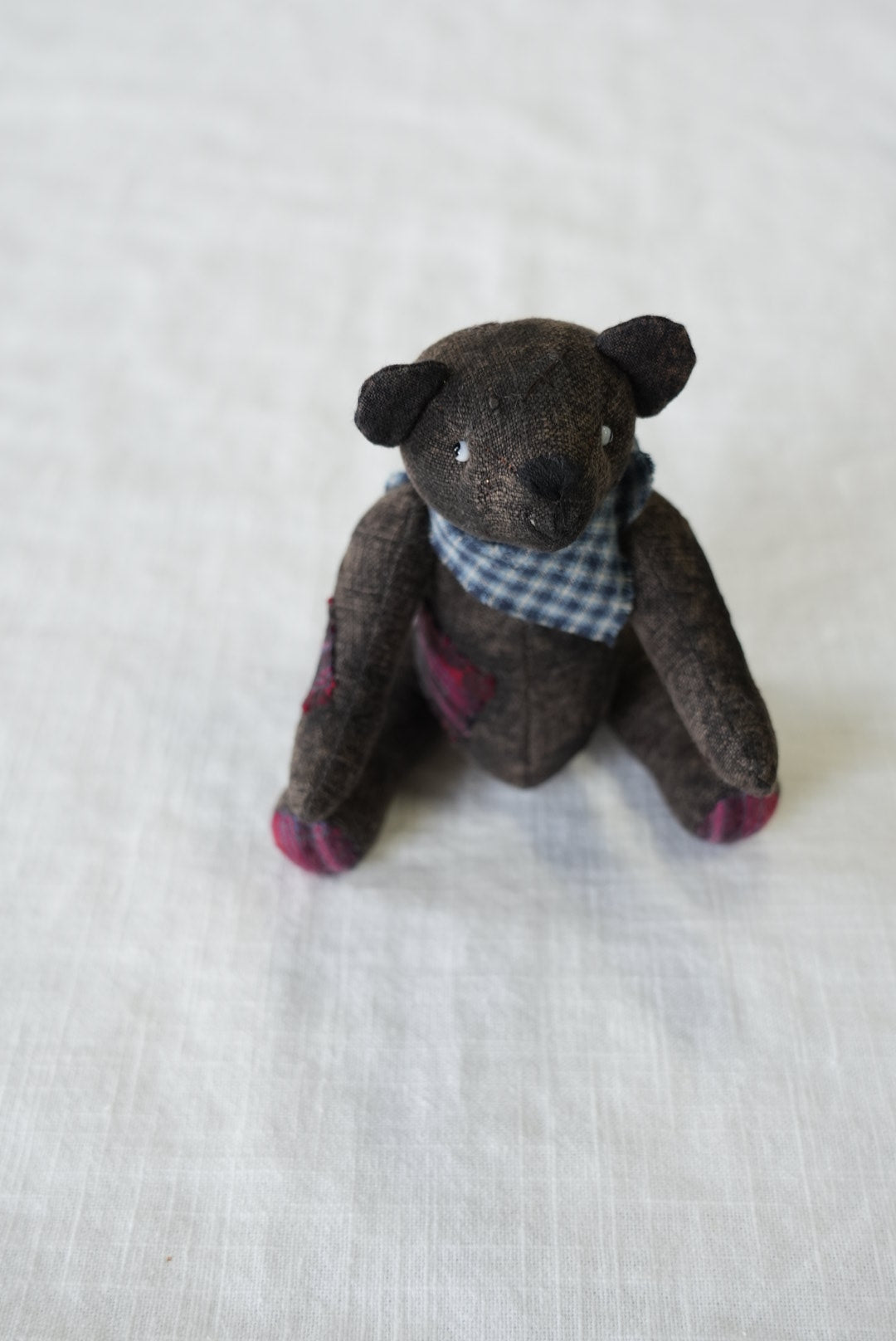 A small handmade fabric teddy bear doll, brown with pink accents on its paws and a blue-and-white checkered shirt, lying on a white textured fabric background.