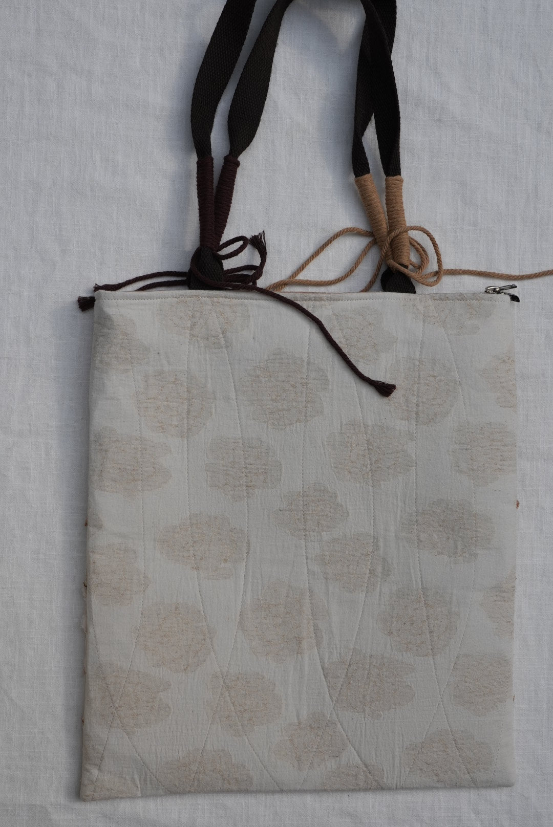 A square fabric cushion or pad with a beige and off-white floral block print pattern, featuring dark brown and tan hanging straps on the left side, laid flat on a white textured fabric background.