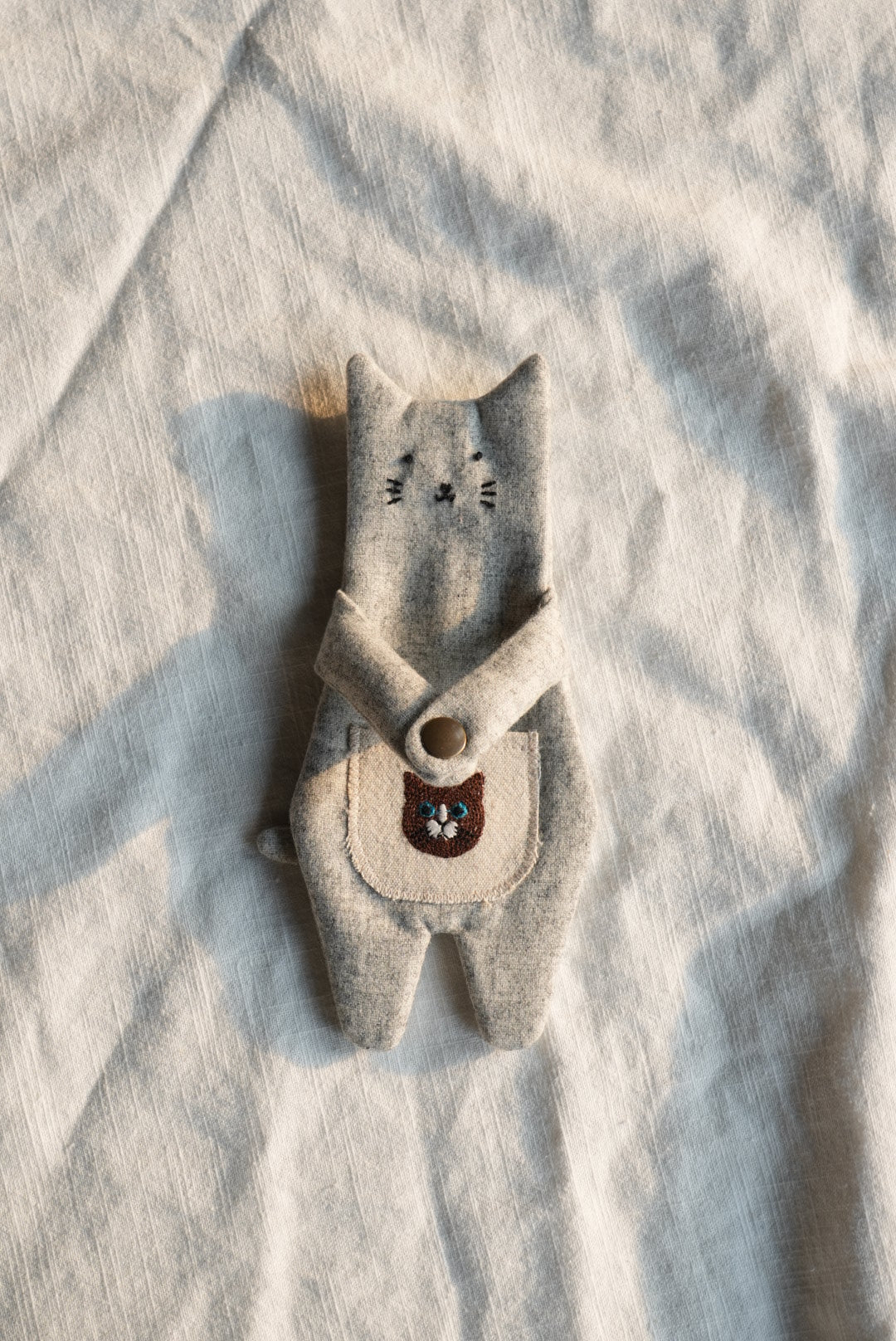 A handmade felted wool toy shaped like a small animal or creature, laid flat on a textured off-white fabric background. The toy is primarily a light oatmeal-beige color with a dark brown embroidered face and small black stitched details.