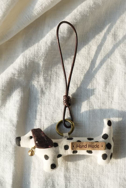 A small bone-shaped dog keychain charm with black spots, a dark brown leather strap, and a small wooden tag that reads 'Hand Made', photographed on a light fabric background.
