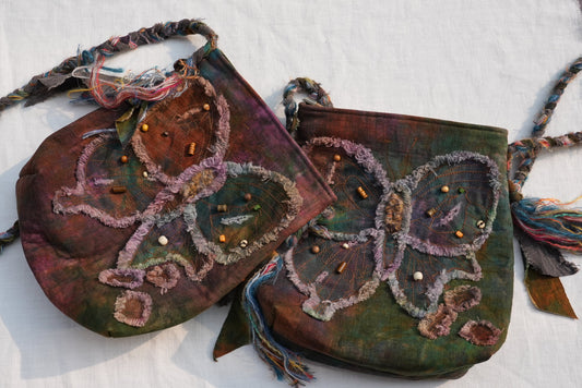 Two handmade butterfly-shaped shoulder bags with braided straps, featuring textured fabric, tassel details, and colorful bead embellishments on a white fabric background.