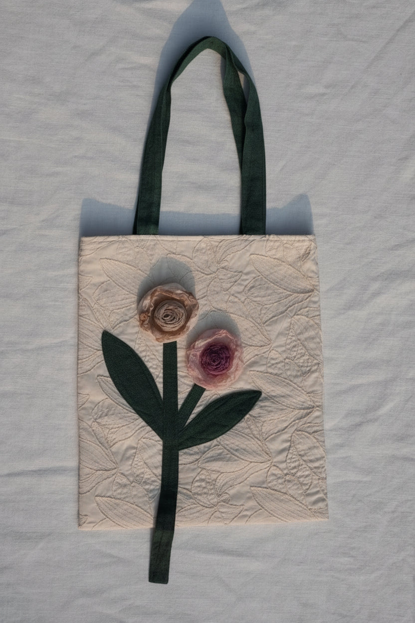 A handmade fabric tote bag with a cream-colored quilted background, featuring a dark green ribbon handle and a stylized embroidered rose design in pink and beige.