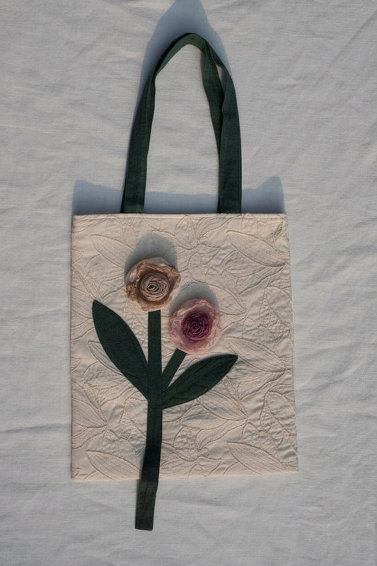 A handmade fabric tote bag with a cream-colored quilted background, featuring a dark green ribbon handle and a stylized embroidered rose design in pink and beige.