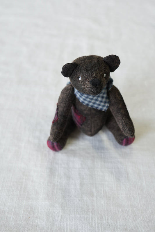A small handmade fabric teddy bear doll, brown with pink accents on its paws and a blue-and-white checkered shirt, lying on a white textured fabric background.