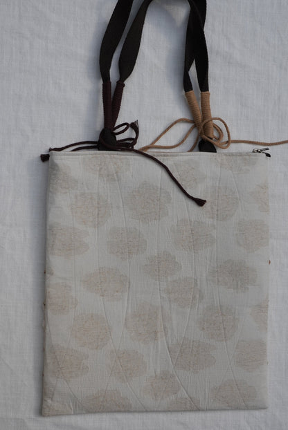 A square fabric cushion or pad with a beige and off-white floral block print pattern, featuring dark brown and tan hanging straps on the left side, laid flat on a white textured fabric background.