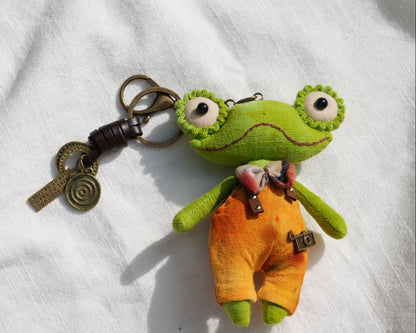 A handmade green frog plush keychain with large embroidered eyes, wearing orange overalls, resting on a white textured fabric background next to a brass keyring and decorative key.