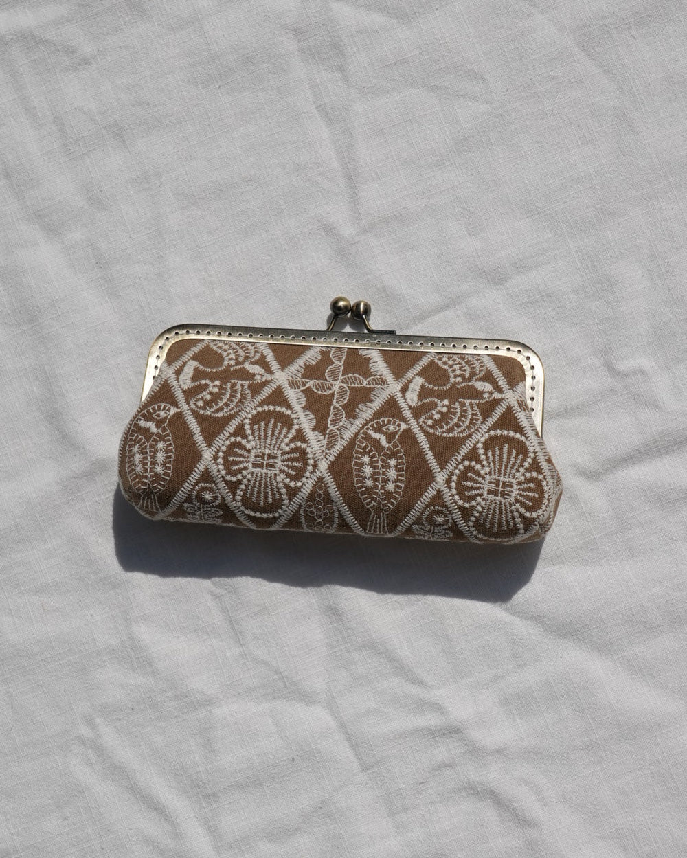 A small rectangular clutch or wallet with a brown base and white geometric floral patterns, featuring a silver-tone metal clasp, resting on a wrinkled white fabric surface.