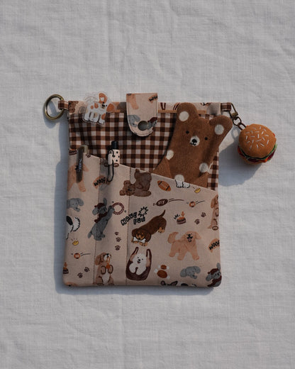A small fabric pouch with a mixed pattern design featuring brown and beige woodland motifs, a brown-and-beige gingham pocket, and a small round macaroni charm, laid flat on a white textured fabric background.