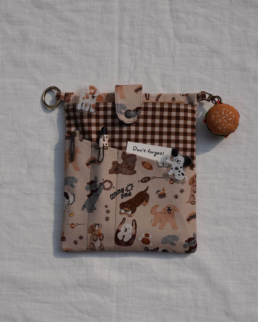 A small fabric pouch with a mixed print design featuring cartoon animals on a beige background and a brown gingham section, shown on a white textured fabric background. It has a metal key ring and a small donut-shaped charm.