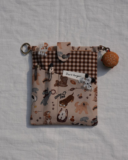 A small fabric pouch with a mixed print design featuring cartoon animals on a beige background and a brown gingham section, shown on a white textured fabric background. It has a metal key ring and a small donut-shaped charm.