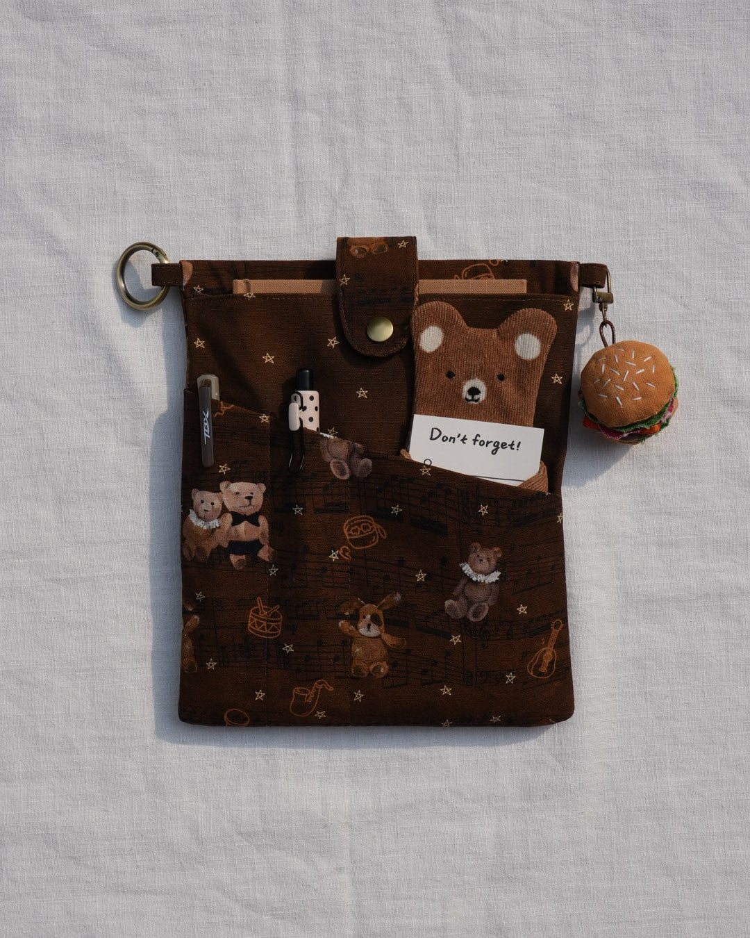 A brown fabric zippered pouch with a teddy bear pattern, featuring a snap-button strap, a small card holder, and a mini hamburger charm, laid flat on a white textured fabric background.