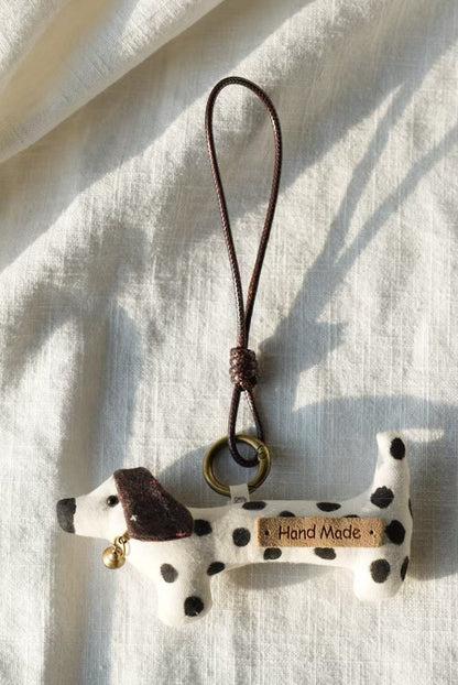 A small bone-shaped dog keychain charm with black spots, a dark brown leather strap, and a small wooden tag that reads 'Hand Made', photographed on a light fabric background.